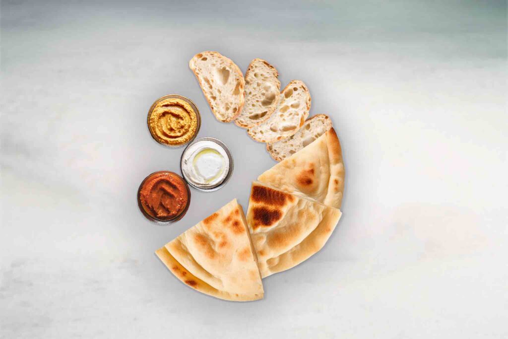 A platter of freshly baked Turkish and Pita bread, served with four colorful dips: spicy capsicum, hummus, eggplant, and tzatziki, ready for sharing.